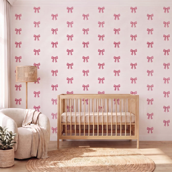 A nursery room scene featuring wallpaper with a repeating pattern of pink bows on a white background, styled with a light wood crib, a cream armchair, a woven floor lamp, and a round jute rug.