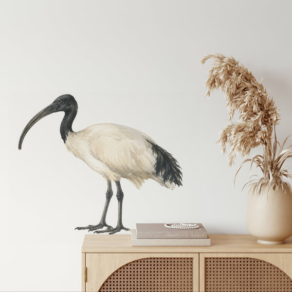 A large wall decal of an African white ibis bird is displayed on a light gray wall above a wooden cabinet, styled with a book and a vase of dried pampas grass.