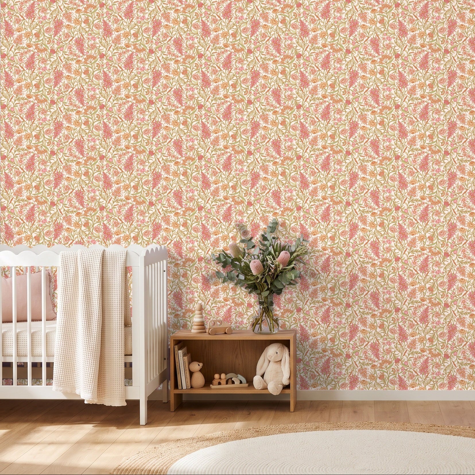 A nursery room featuring a pink and yellow floral patterned wallpaper on the wall, styled with a white crib, a wooden side table with flowers, a white dresser, and a round rug on a light wood floor.