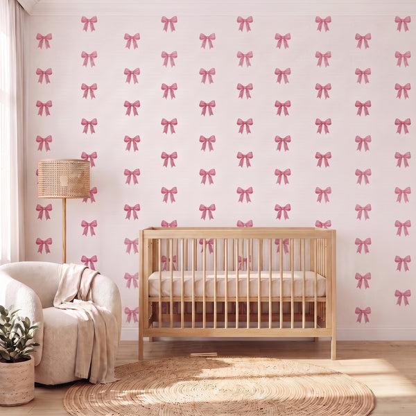 A nursery room scene featuring wallpaper with a repeating pattern of pink bows on a white background, styled with a light wood crib, a cream armchair, a woven floor lamp, and a round jute rug.