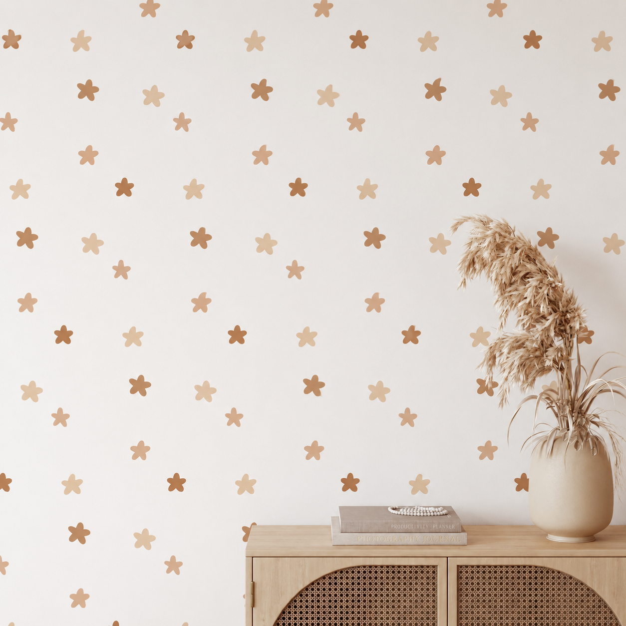 A styled interior scene showing a cream wall with a repeating pattern of small metallic gold and bronze star stickers, above a light wood cabinet with a vase of dried grass.
