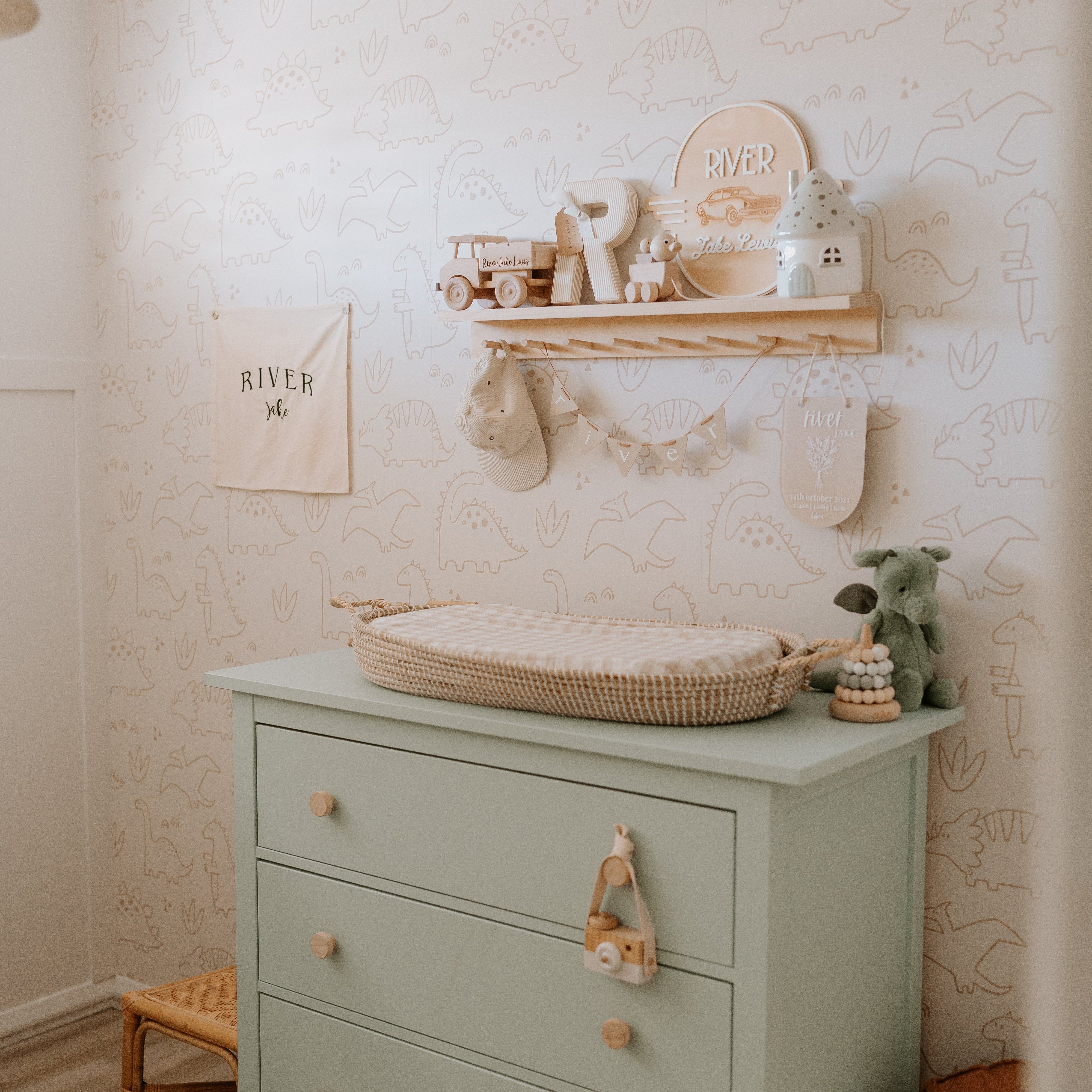 A styled nursery corner featuring a sage green three-drawer dresser with wooden knobs, topped with a woven changing tray. The room has pale blush wallpaper with subtle animal line drawings, a wooden floating shelf with nursery decor, and a small wooden stool.