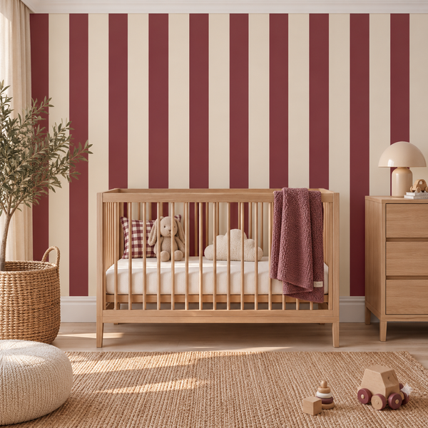 A styled nursery room featuring a light wood crib with a white mattress and bedding, set against a wall with vertical burgundy and cream stripes. A matching wooden dresser with a lamp is on the right, and a woven basket, potted plant, and round pouf are on the left.