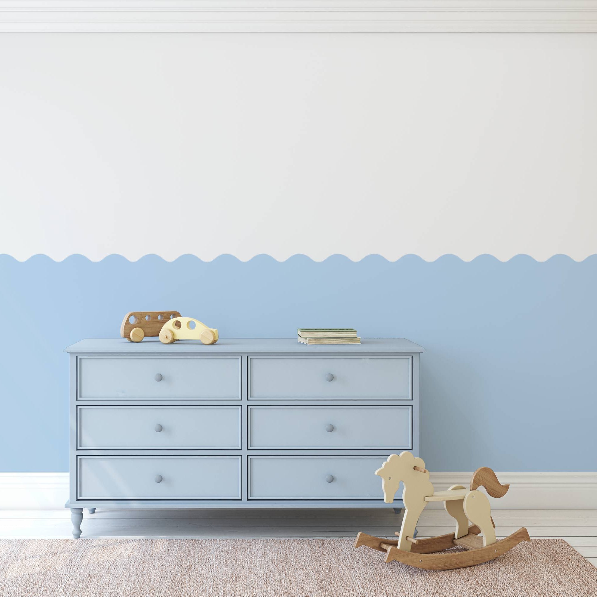 A styled nursery room scene featuring a light blue dresser against a white and light blue scalloped wall, with a wooden rocking horse and a small toy car on top.