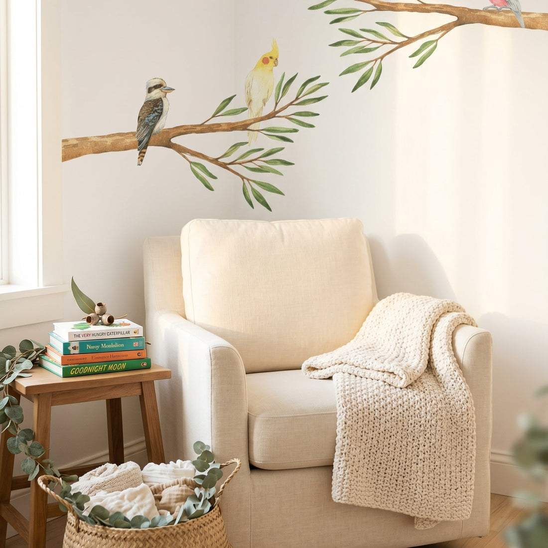 Beige armchair with a blanket, small table with books, and wall decal of birds on branches in a cozy room.