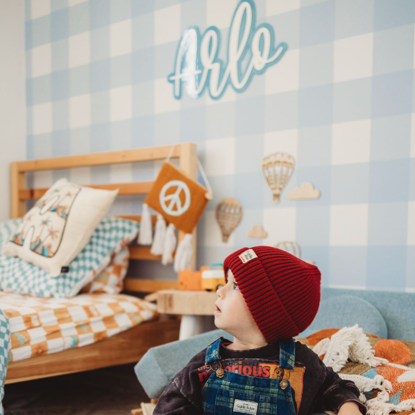 Child in a bedroom with checkered walls, wooden furniture, and toys on the floor.