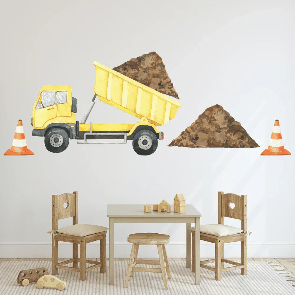 A wall decal of a yellow dump truck with a load of dirt, accompanied by two orange safety cones, applied on a white wall in a children's room setting with wooden furniture and toys.