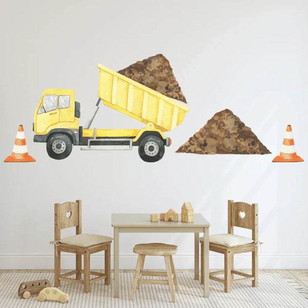 A wall decal of a yellow dump truck with a load of dirt, accompanied by two orange safety cones, applied on a white wall in a children's room setting with wooden furniture and toys.