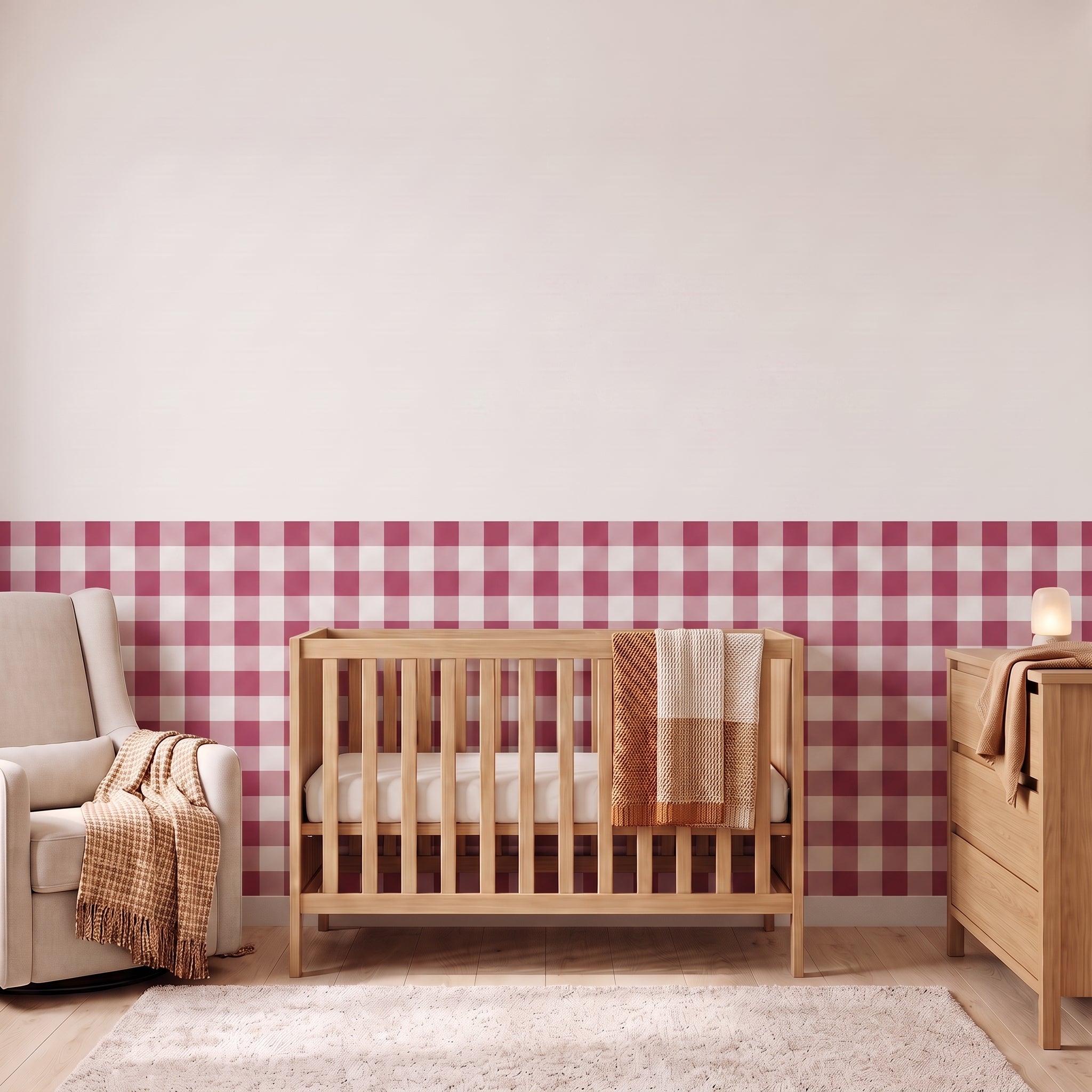 A styled nursery with berry pink gingham check wallpaper across the lower half of the wall. The room has a light wood crib with white bedding and a berry and tan blanket, a cream armchair with a woven throw, a matching wood dresser with a lamp, and a soft rug on a light wood floor.