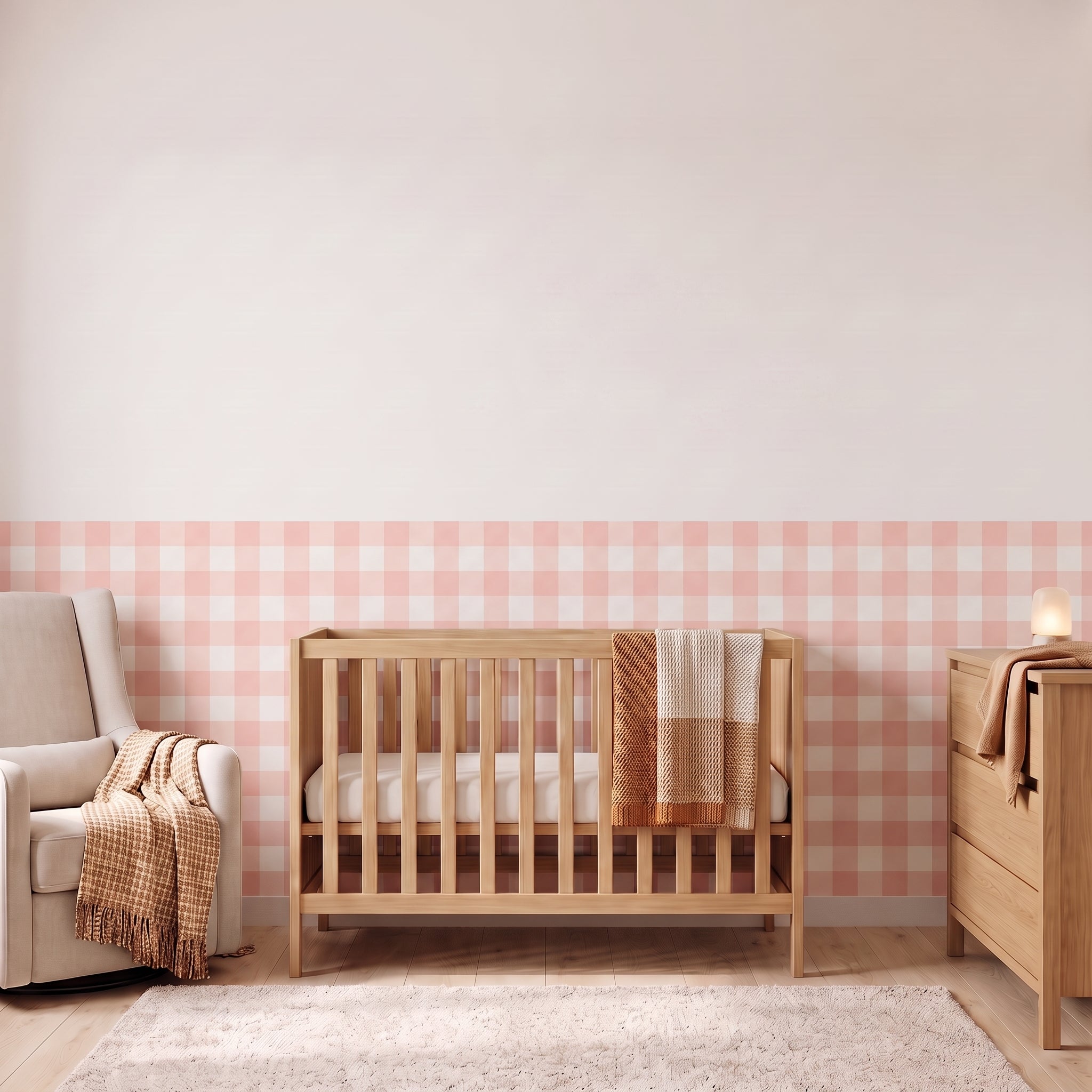 A styled nursery with blush pink gingham check wallpaper across the lower half of the wall. The room has a light wood crib with white bedding and a blush and tan blanket, a cream armchair with a woven throw, a matching wood dresser with a lamp, and a soft rug on a light wood floor.