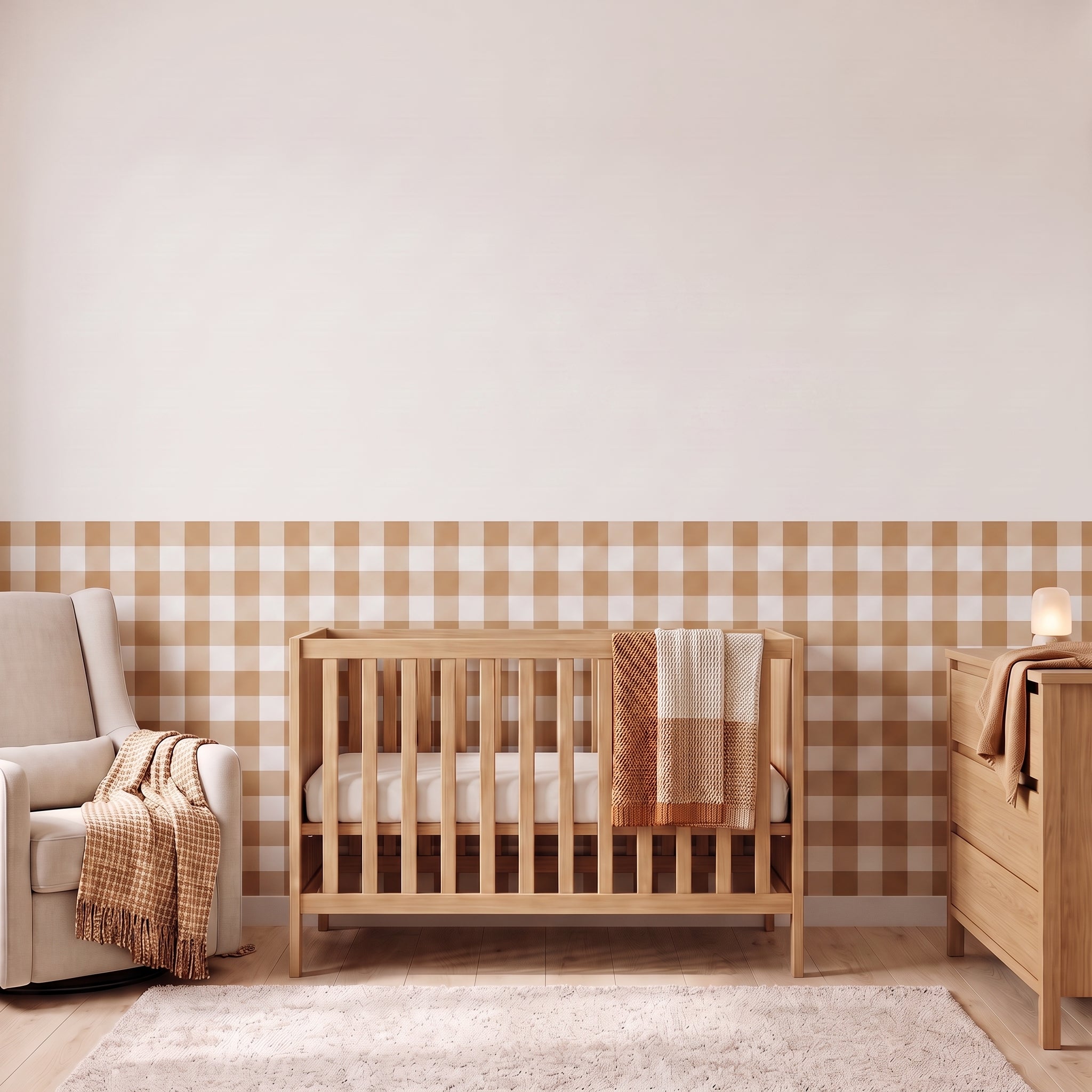A styled nursery with camel tan gingham check wallpaper across the lower half of the wall. The room has a light wood crib with white bedding and a camel and cream checked blanket, a cream armchair with a camel and cream fringed throw, a matching light wood dresser with a lamp, and a soft cream rug on a light wood floor.