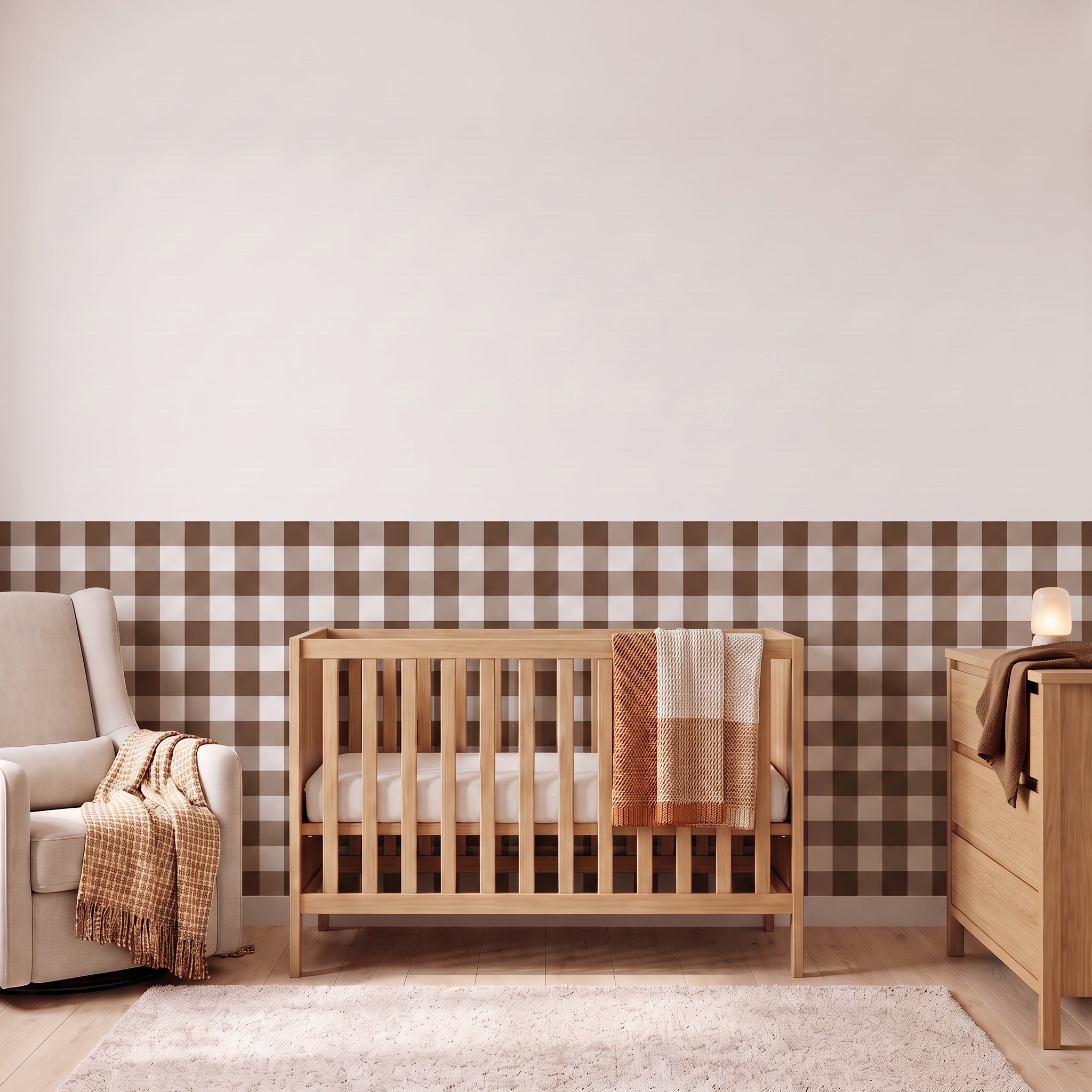 A styled nursery with chocolate brown gingham check wallpaper across the lower half of the wall. The room has a light wood crib with white bedding and a brown and cream checked blanket, a cream armchair with a brown and cream fringed throw, a matching light wood dresser with a lamp, and a soft cream rug on a light wood floor.