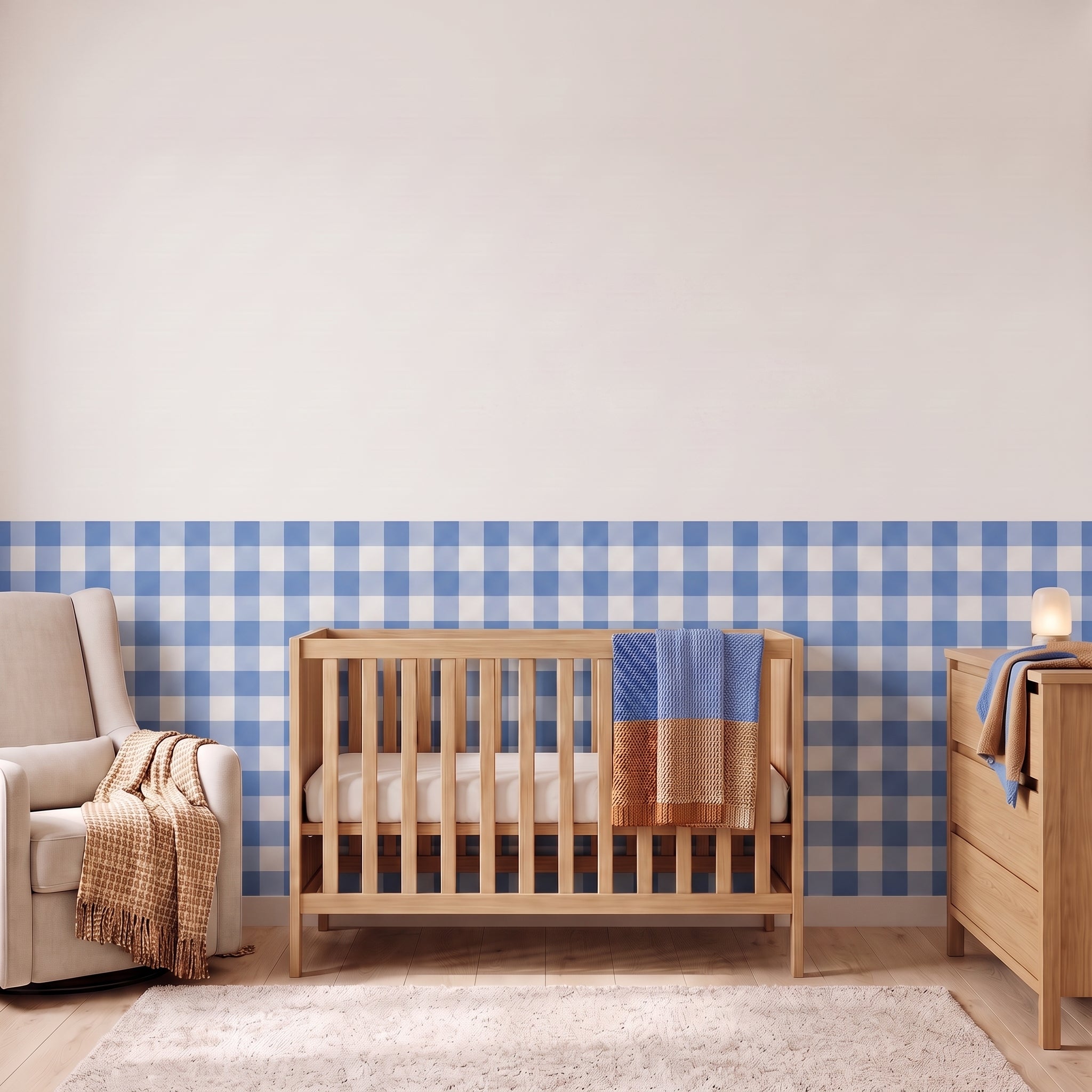 A styled nursery with cornflower blue gingham check wallpaper across the lower half of the wall. The room has a light wood crib with white bedding and a cornflower blue and tan blanket, a cream armchair with a woven throw, a matching wood dresser with a lamp, and a soft rug on a light wood floor.