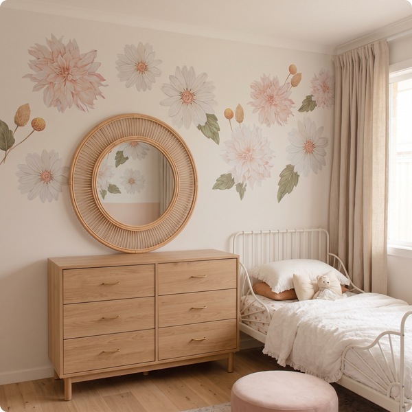 A styled children's bedroom featuring a light wood six-drawer dresser with a round woven rattan mirror, a single bed with white bedding, and a pink blush velvet ottoman. The wall is decorated with large pastel pink and white dahlia floral decals.