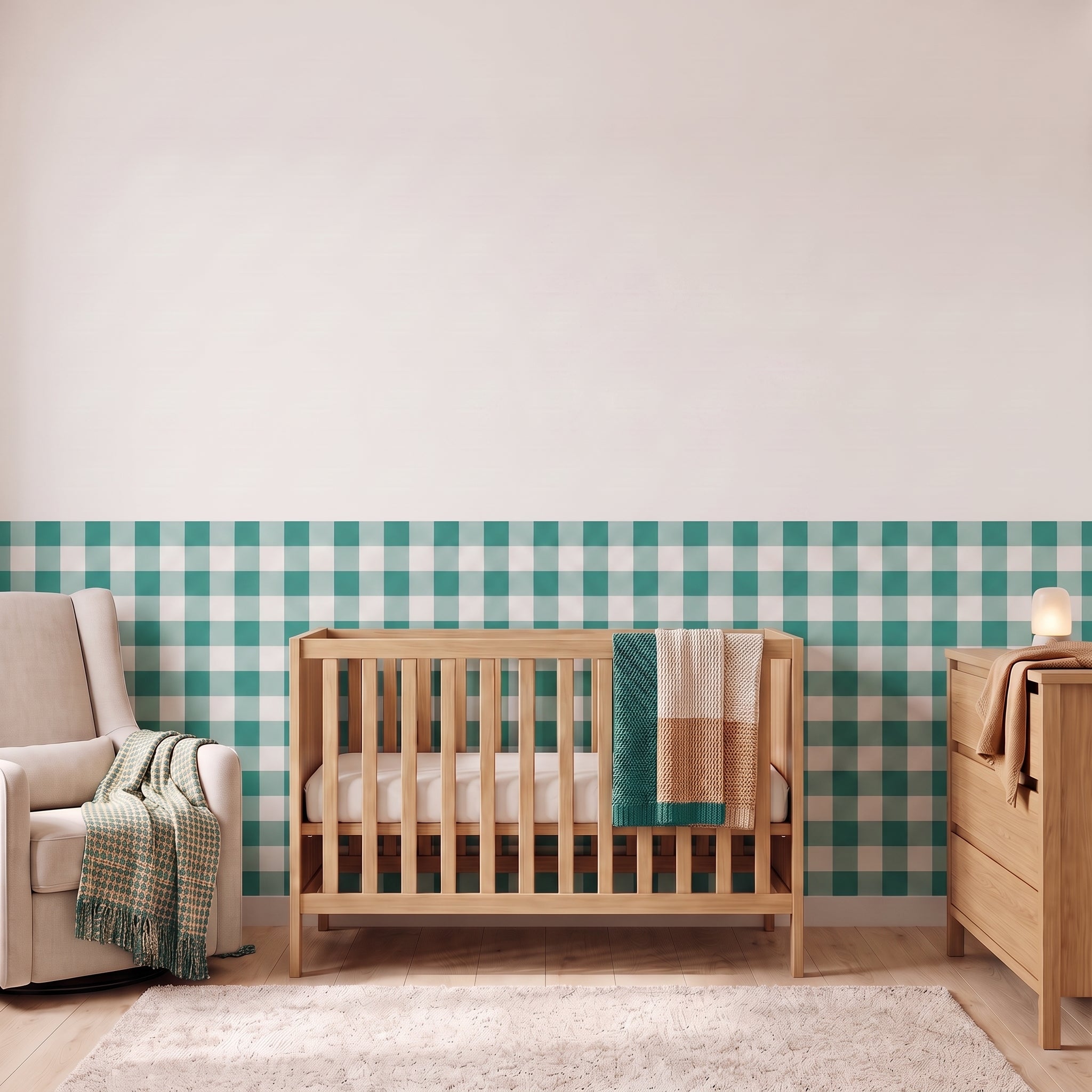 A styled nursery with deep teal gingham check wallpaper across the lower half of the wall. The room has a light wood crib with white bedding and a teal and tan blanket, a cream armchair with a woven throw, a matching wood dresser with a lamp, and a soft rug on a light wood floor.