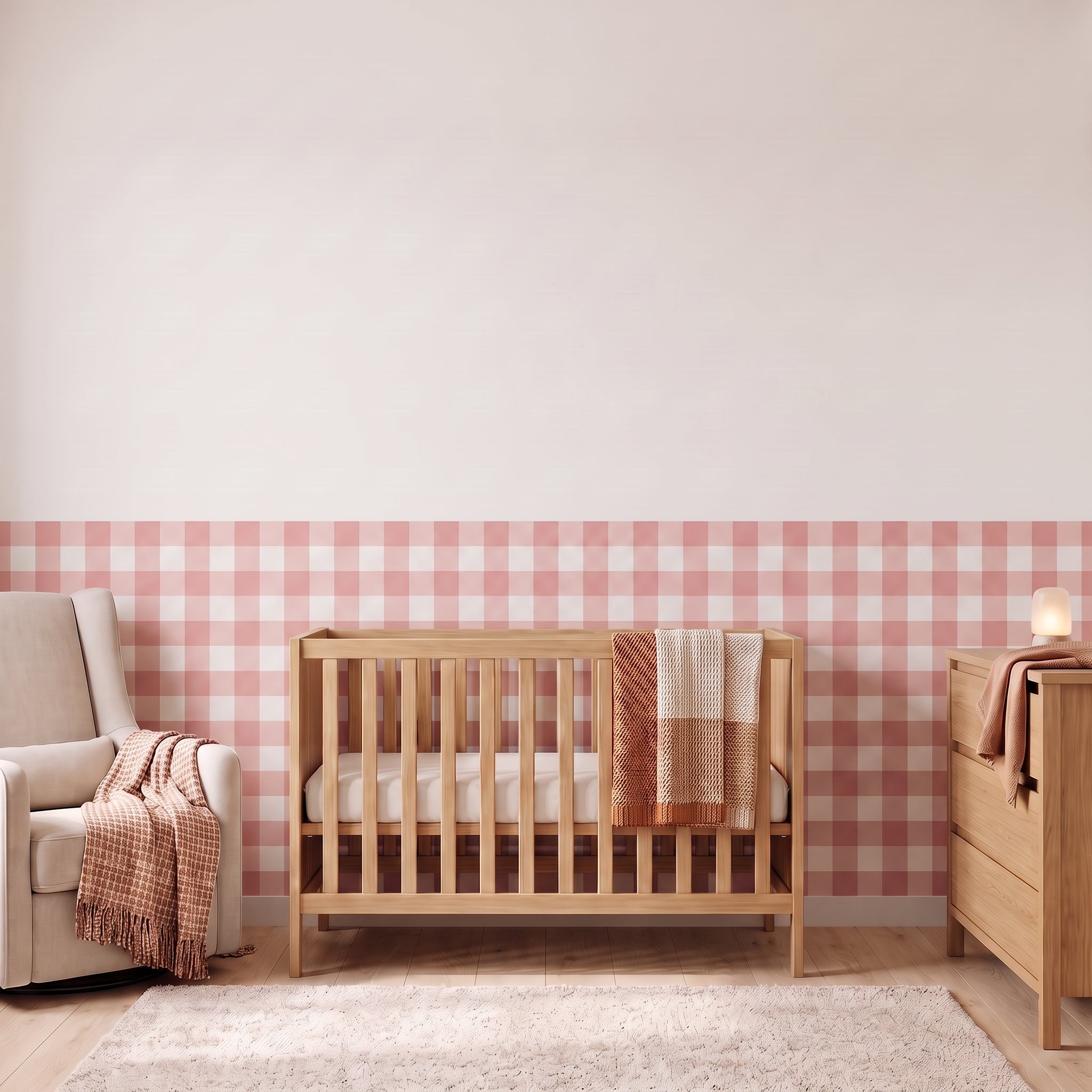 A styled nursery with dusty rose gingham check wallpaper across the lower half of the wall. The room has a light wood crib with white bedding and a dusty rose and tan blanket, a cream armchair with a woven throw, a matching wood dresser with a lamp, and a soft rug on a light wood floor.