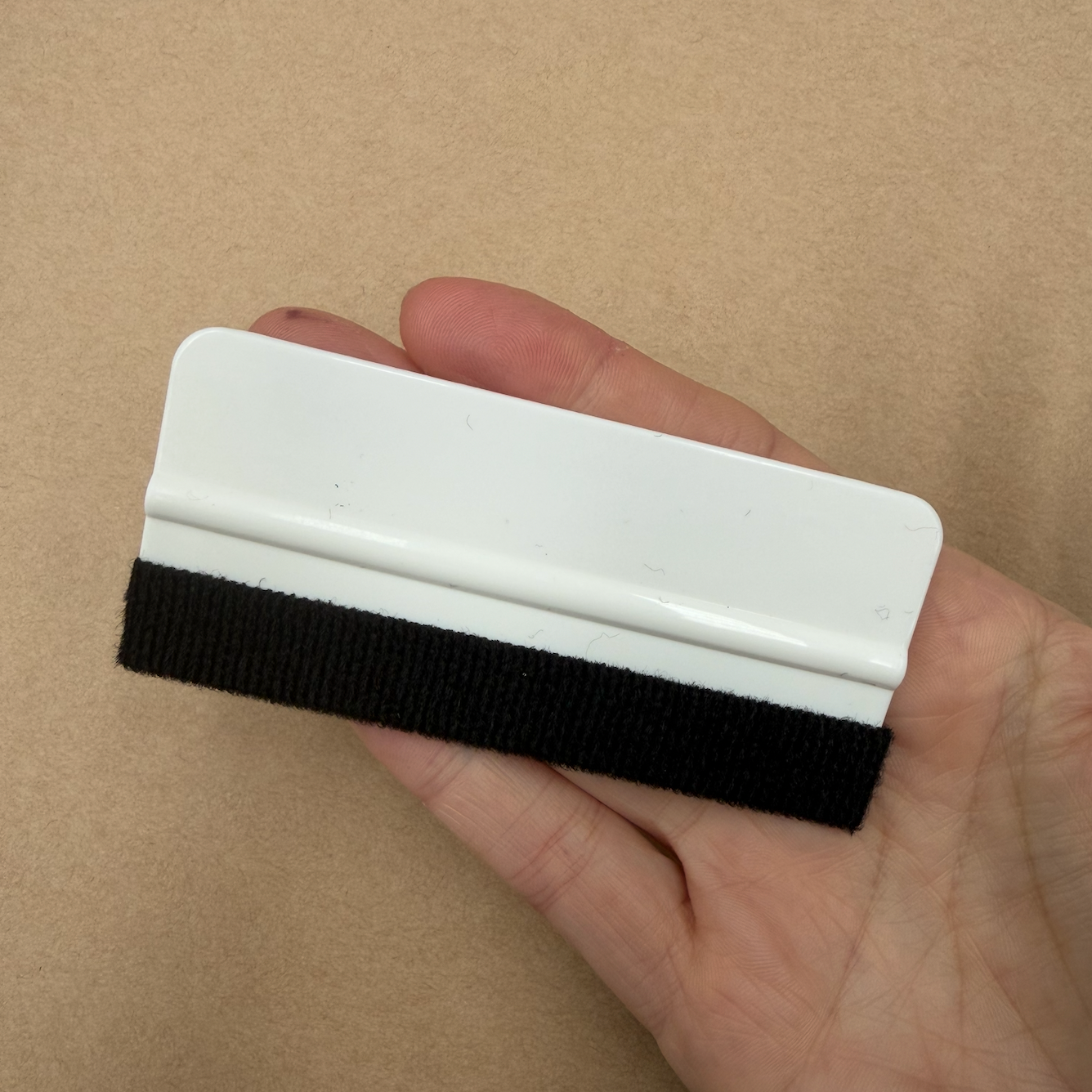 A hand holding a small white plastic scraper or squeegee with a black textured rubber or fabric strip along the bottom edge, set against a plain tan background.
