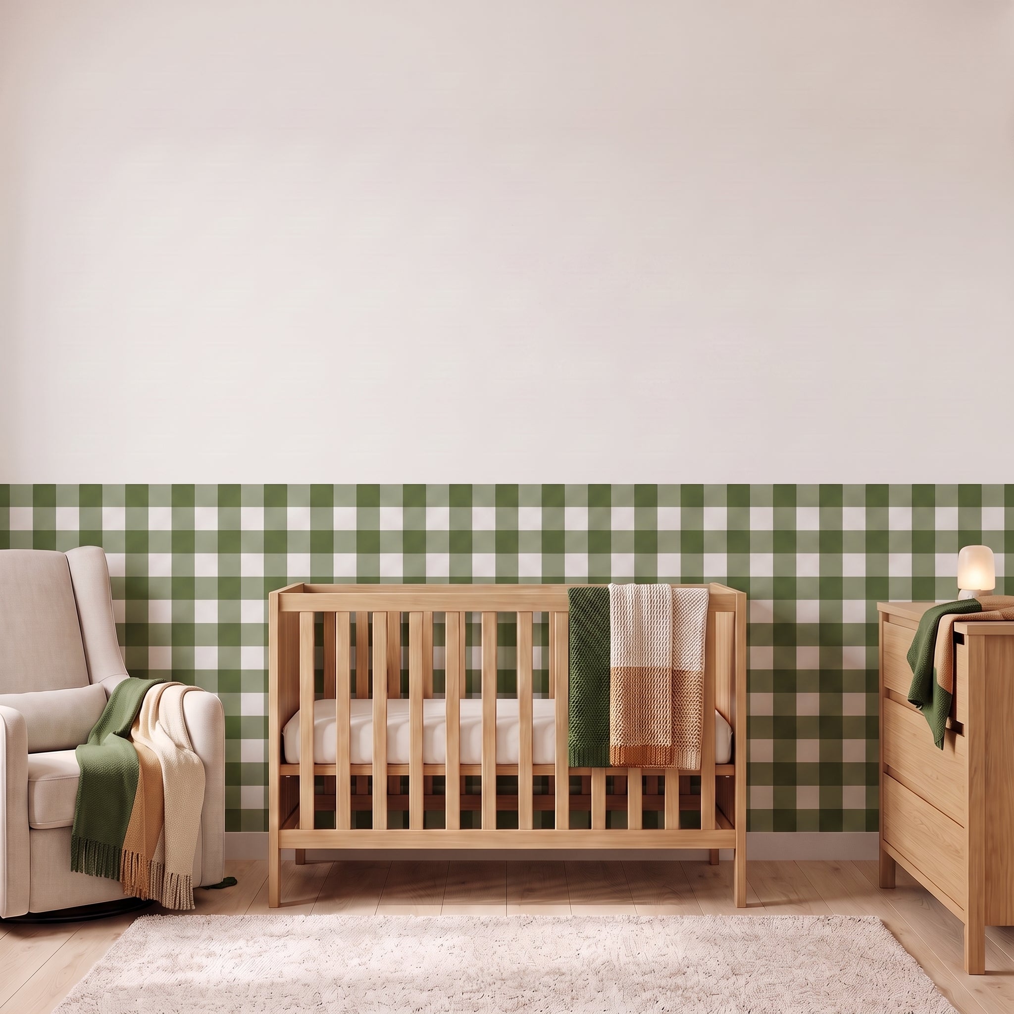 A styled nursery with forest green gingham check wallpaper across the lower half of the wall. The room has a light wood crib with white bedding and a forest green and cream checked blanket, a cream armchair with a green and cream fringed throw, a matching light wood dresser with a lamp, and a soft cream rug on a light wood floor.