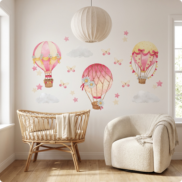 A styled nursery with pink and yellow hot air balloon wall decals, clouds, stars and butterflies on a light wall, furnished with a rattan bassinet, a cream boucle armchair with a throw, a hanging paper lantern and a light wood floor.
