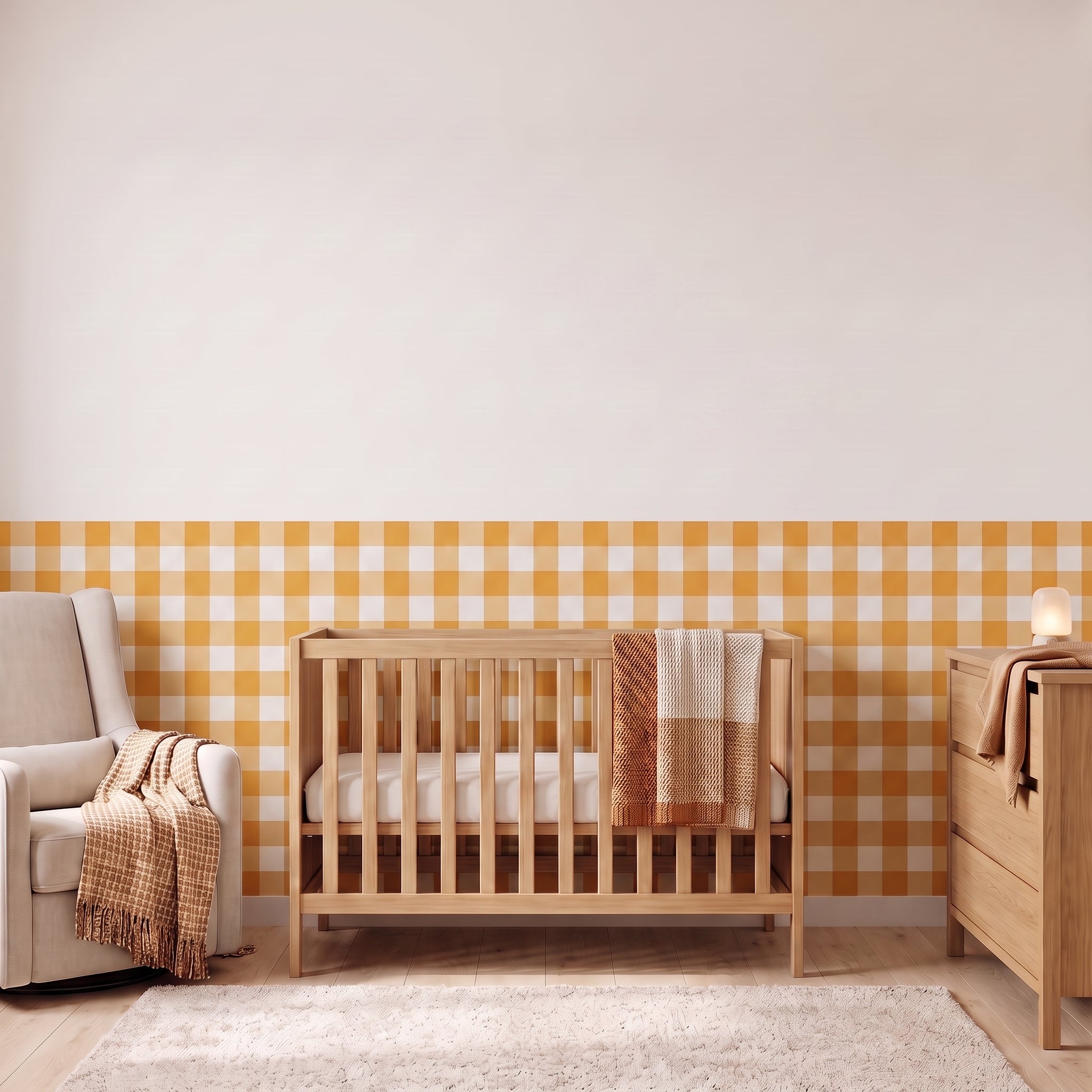 A styled nursery with marigold yellow gingham check wallpaper across the lower half of the wall. The room has a light wood crib with white bedding and a marigold and cream checked blanket, a cream armchair with a marigold and cream fringed throw, a matching light wood dresser with a lamp, and a soft cream rug on a light wood floor.