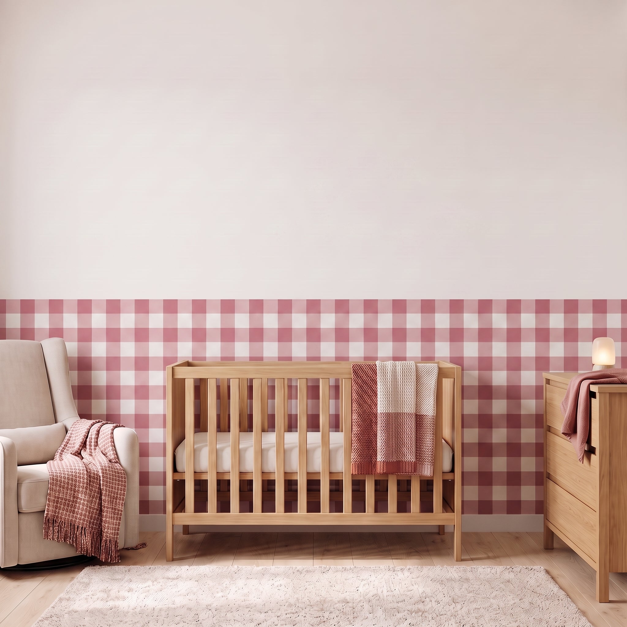 A styled nursery with mauve rose gingham check wallpaper across the lower half of the wall. The room has a light wood crib with white bedding and a mauve-rose and cream blanket, a cream armchair with a mauve-rose woven throw, a matching wood dresser with a lamp, and a soft rug on a light wood floor.