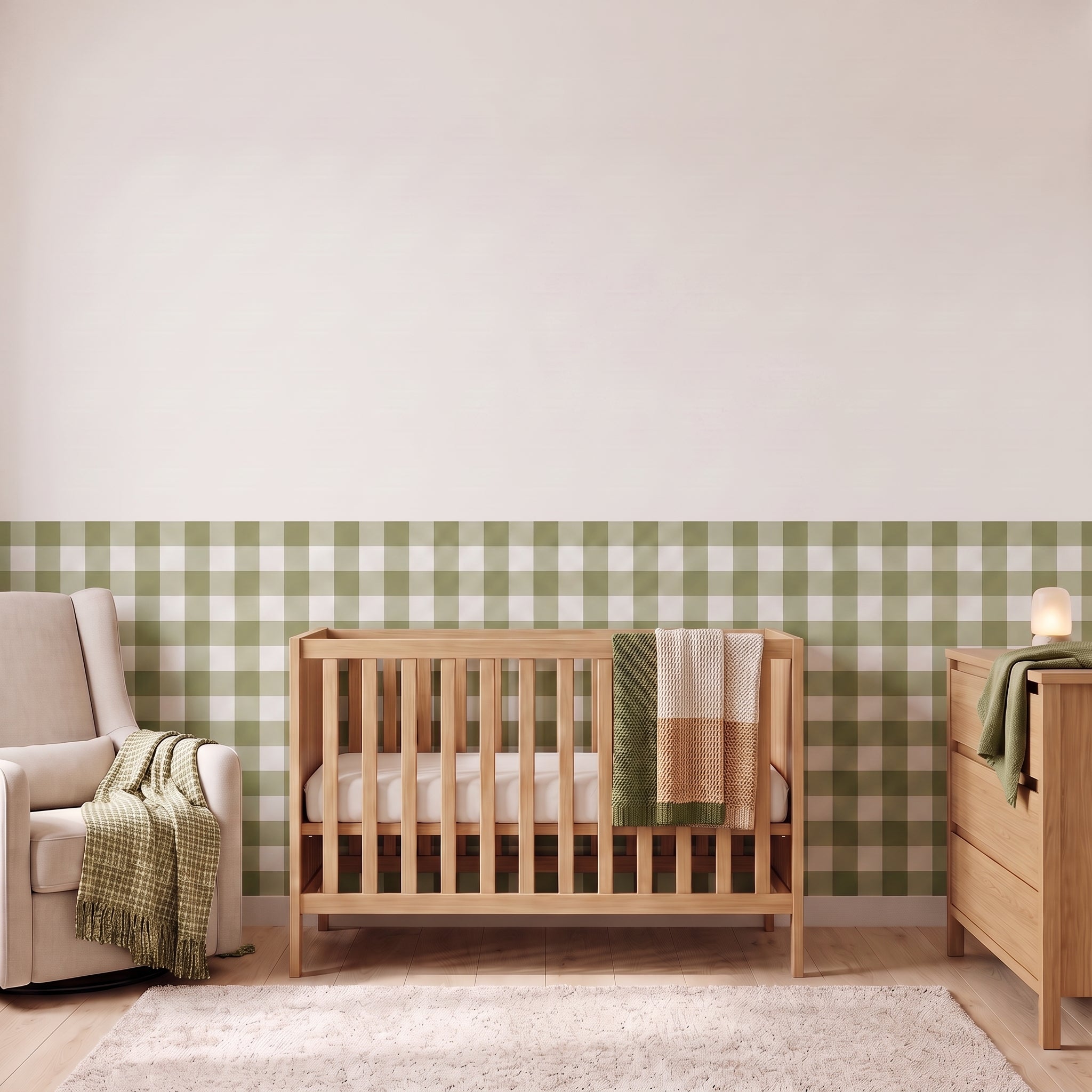 A styled nursery with moss green gingham check wallpaper across the lower half of the wall. The room has a light wood crib with white bedding and a moss green and cream checked blanket, a cream armchair with a green and cream fringed throw, a matching light wood dresser with a lamp, and a soft cream rug on a light wood floor.