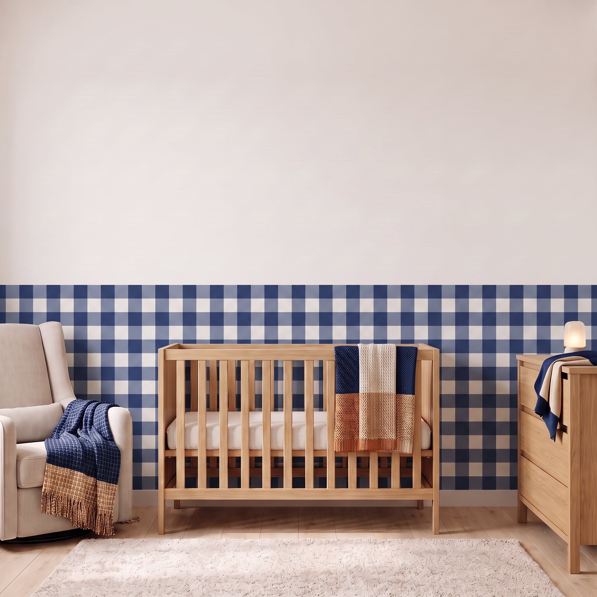 A styled nursery with navy gingham check wallpaper across the lower half of the wall. The room has a light wood crib with white bedding and a navy and tan blanket, a cream armchair with a navy throw, a matching wood dresser with a lamp, and a soft rug on a light wood floor.