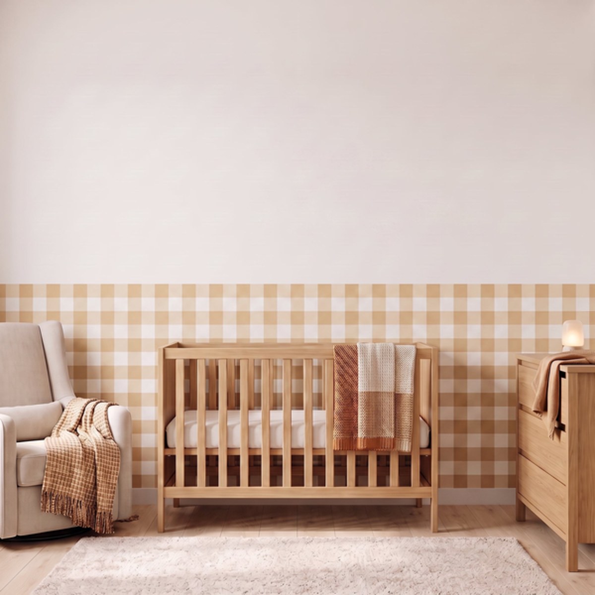 A styled nursery with pale oatmeal beige gingham check wallpaper across the lower half of the wall. The room has a light wood crib with white bedding and an oat and cream checked blanket, a cream armchair with an oat and cream fringed throw, a matching light wood dresser with a lamp, and a soft cream rug on a light wood floor.