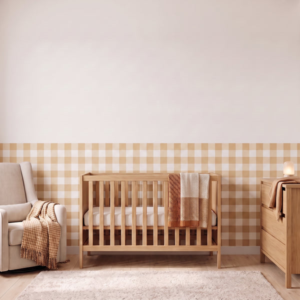 A styled nursery with pale oatmeal beige gingham check wallpaper across the lower half of the wall. The room has a light wood crib with white bedding and an oat and cream checked blanket, a cream armchair with an oat and cream fringed throw, a matching light wood dresser with a lamp, and a soft cream rug on a light wood floor.