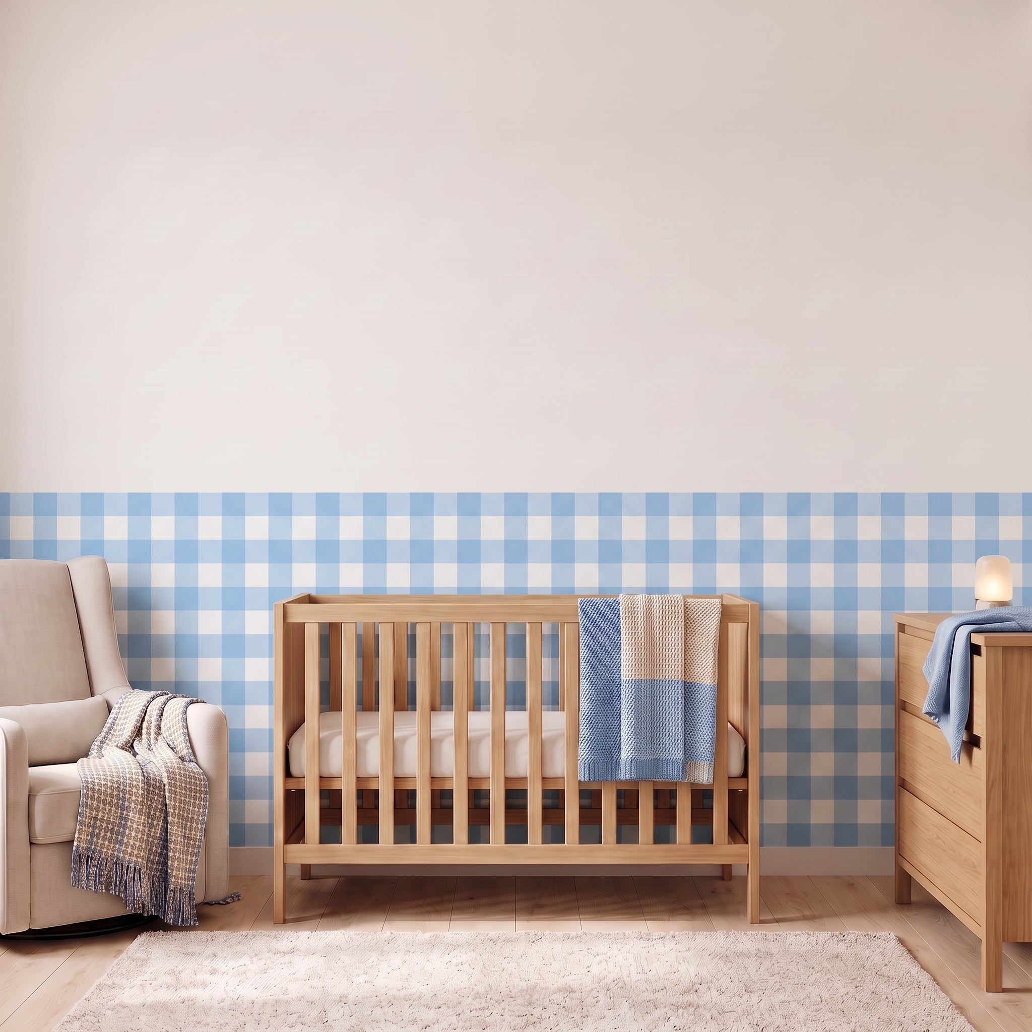 A styled nursery with powder blue gingham check wallpaper across the lower half of the wall. The room has a light wood crib with white bedding and a powder blue and tan blanket, a cream armchair with a woven throw, a matching wood dresser with a lamp, and a soft rug on a light wood floor.