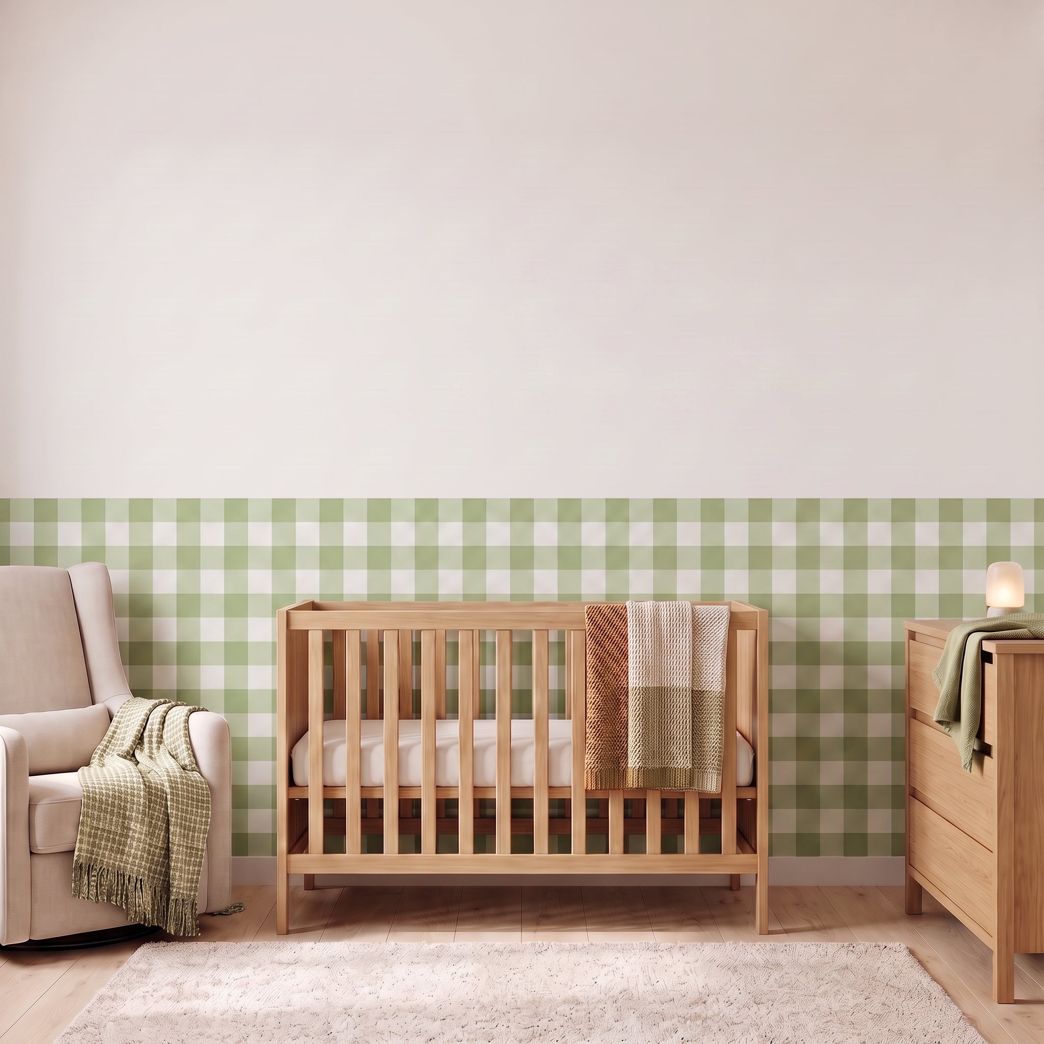 A styled nursery with sage green gingham check wallpaper across the lower half of the wall. The room has a light wood crib with white bedding and a sage green and cream checked blanket, a cream armchair with a green and cream fringed throw, a matching light wood dresser with a lamp, and a soft cream rug on a light wood floor.
