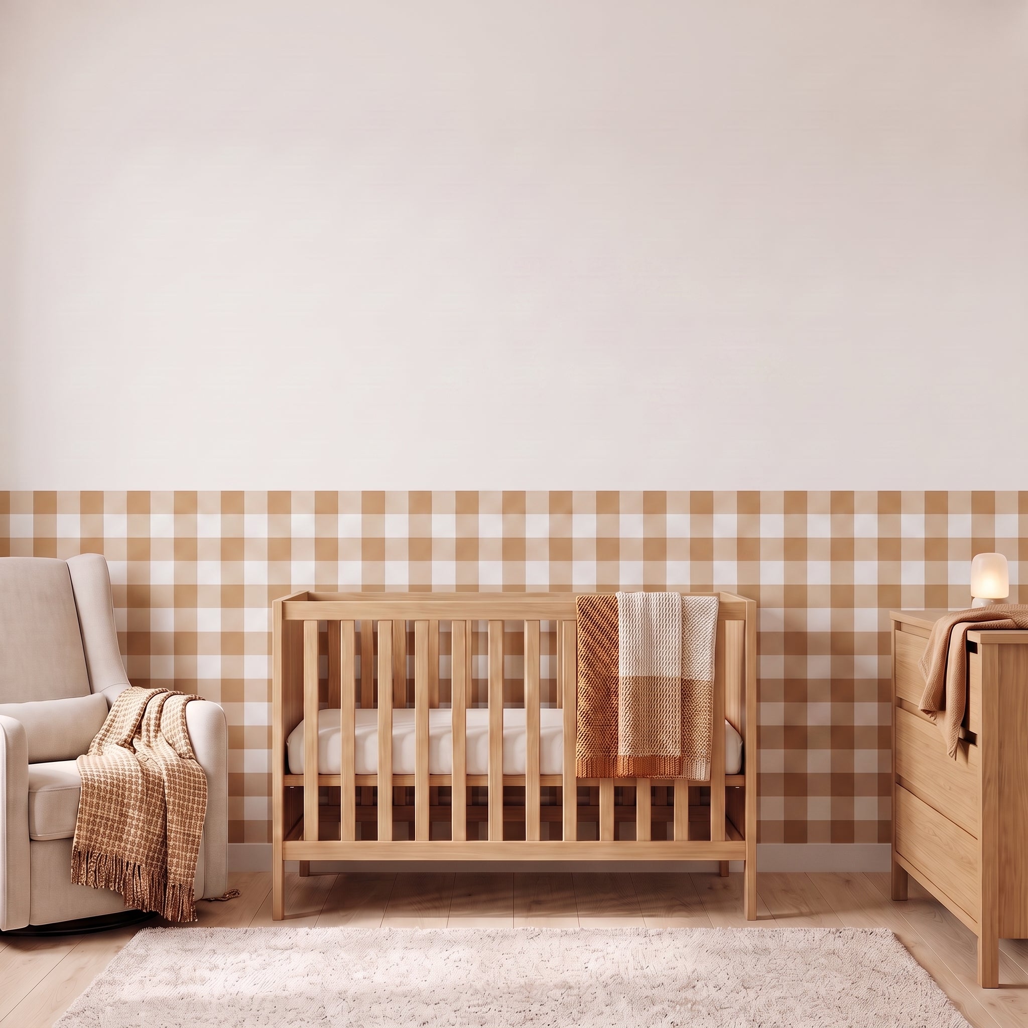 A styled nursery with sandy beige gingham check wallpaper across the lower half of the wall. The room has a light wood crib with white bedding and a sand and cream checked blanket, a cream armchair with a sand and cream fringed throw, a matching light wood dresser with a lamp, and a soft cream rug on a light wood floor.