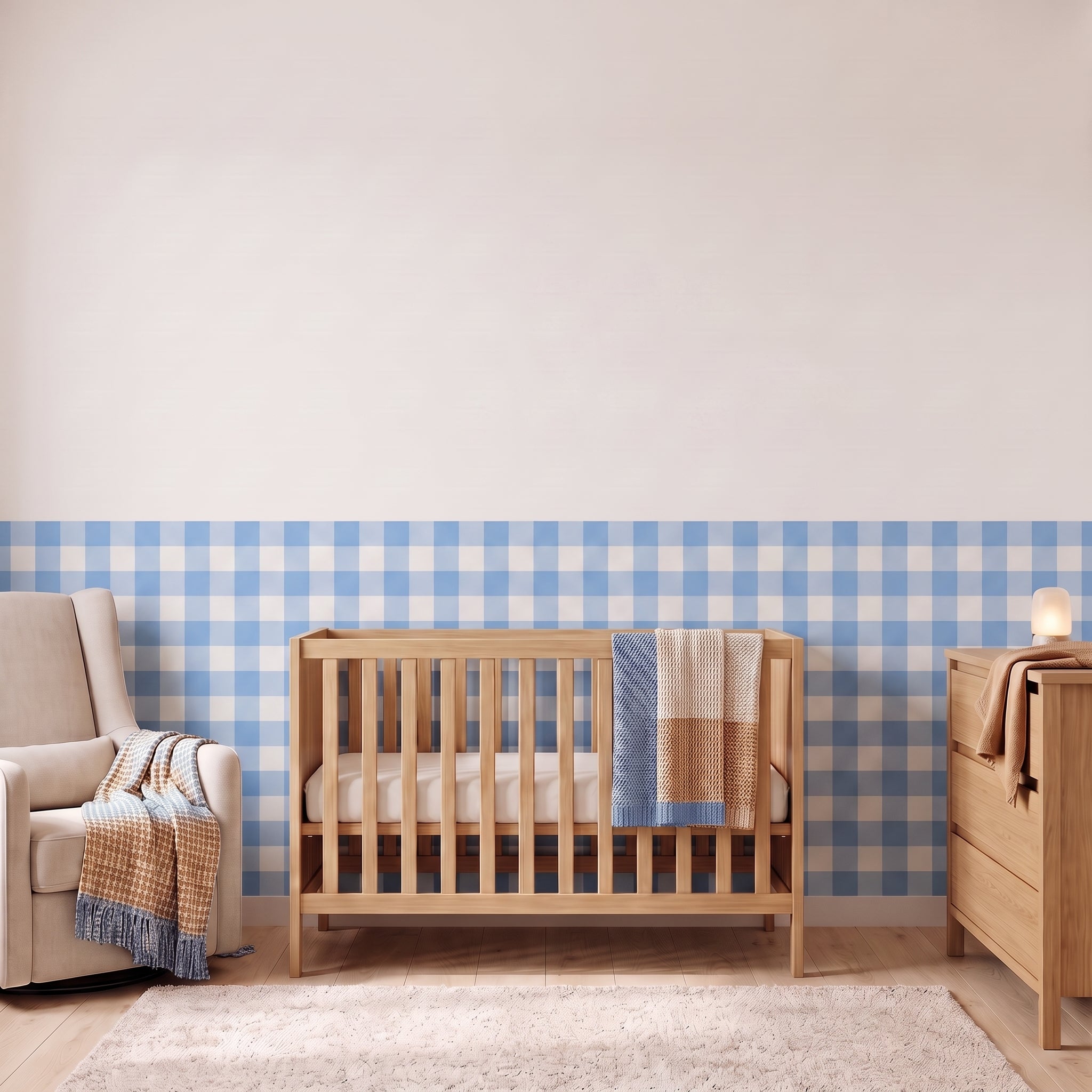 A styled nursery with sky blue gingham check wallpaper across the lower half of the wall. The room has a light wood crib with white bedding and a sky blue and tan blanket, a cream armchair with a woven throw, a matching wood dresser with a lamp, and a soft rug on a light wood floor.
