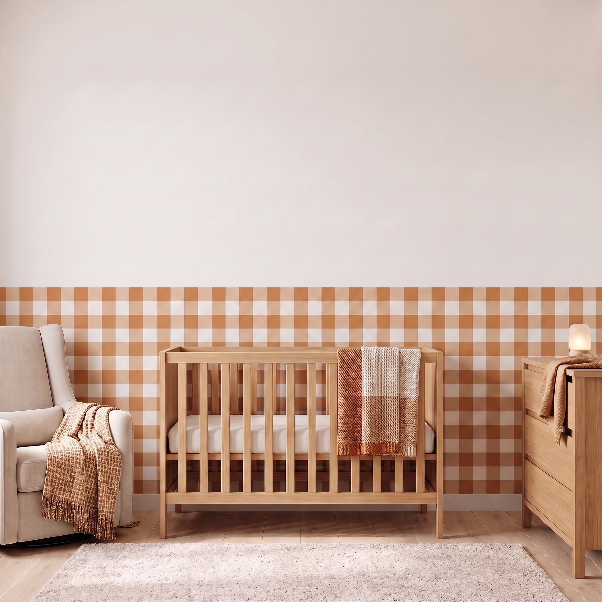 A styled nursery with terracotta gingham check wallpaper across the lower half of the wall. The room has a light wood crib with white bedding and a terracotta and cream checked blanket, a cream armchair with a terracotta and cream fringed throw, a matching light wood dresser with a lamp, and a soft cream rug on a light wood floor.