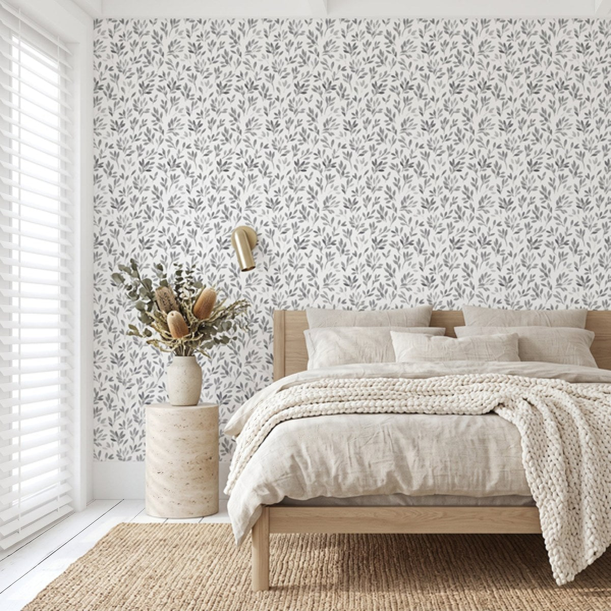 A styled bedroom interior featuring a white wall covered in a delicate gray botanical leaf pattern wallpaper. The room is styled with a light wood platform bed, neutral beige linens, a textured cream knit throw blanket, a rustic wood side table, a vase with dried botanicals, and a gold wall sconce.