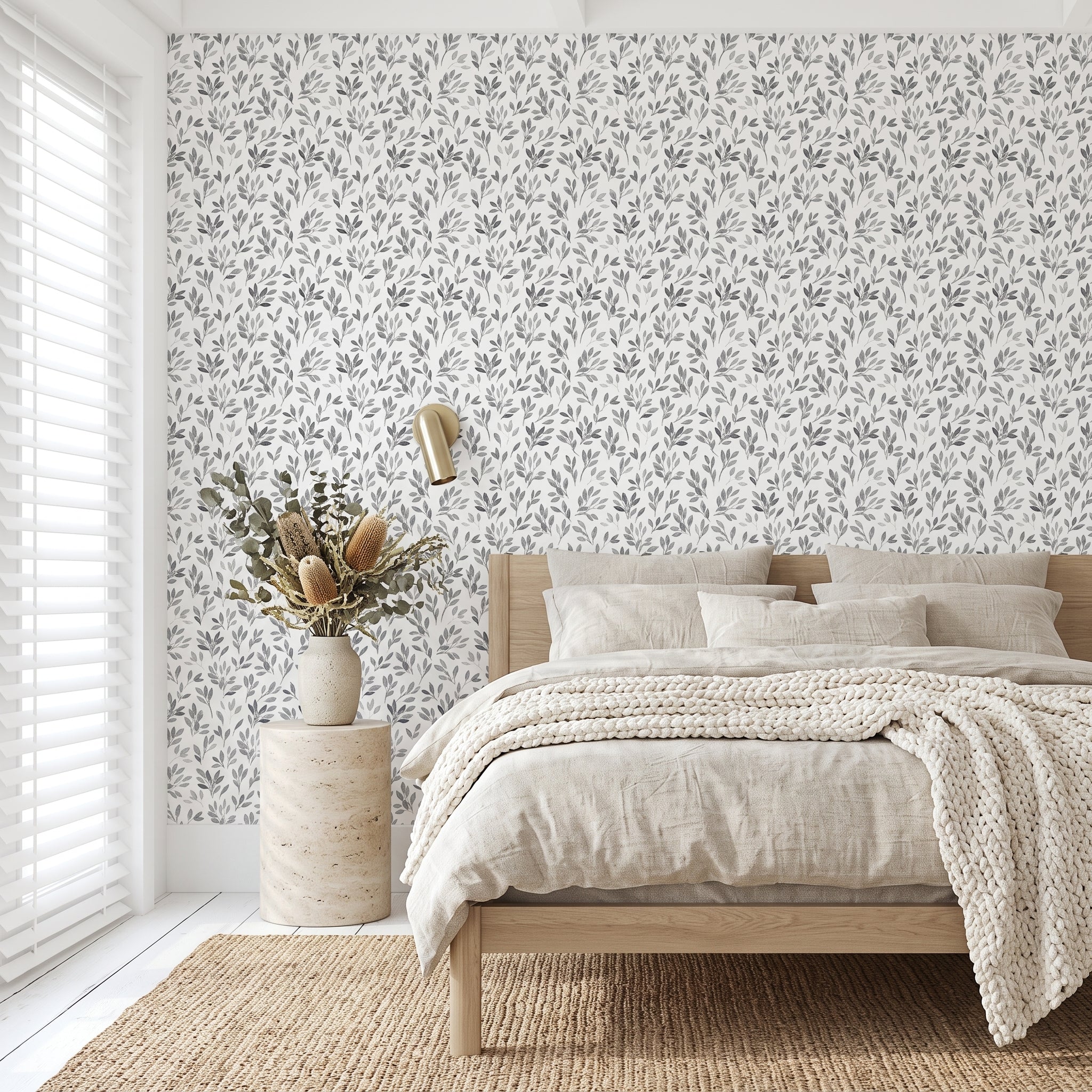 A styled bedroom interior featuring a white wall covered in a delicate gray botanical leaf pattern wallpaper. The room is styled with a light wood platform bed, neutral beige linens, a textured cream knit throw blanket, a rustic wood side table, a vase with dried botanicals, and a gold wall sconce.
