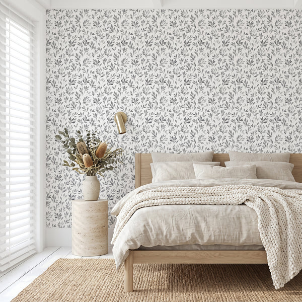 A styled bedroom interior featuring a white wall covered in a delicate gray botanical leaf pattern wallpaper. The room is styled with a light wood platform bed, neutral beige linens, a textured cream knit throw blanket, a rustic wood side table, a vase with dried botanicals, and a gold wall sconce.