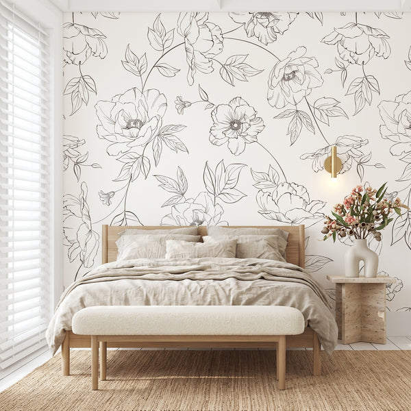 A styled bedroom featuring a white wallpaper with elegant black floral line art, a light wood bed with beige bedding, a matching upholstered bench, and a side table with flowers.